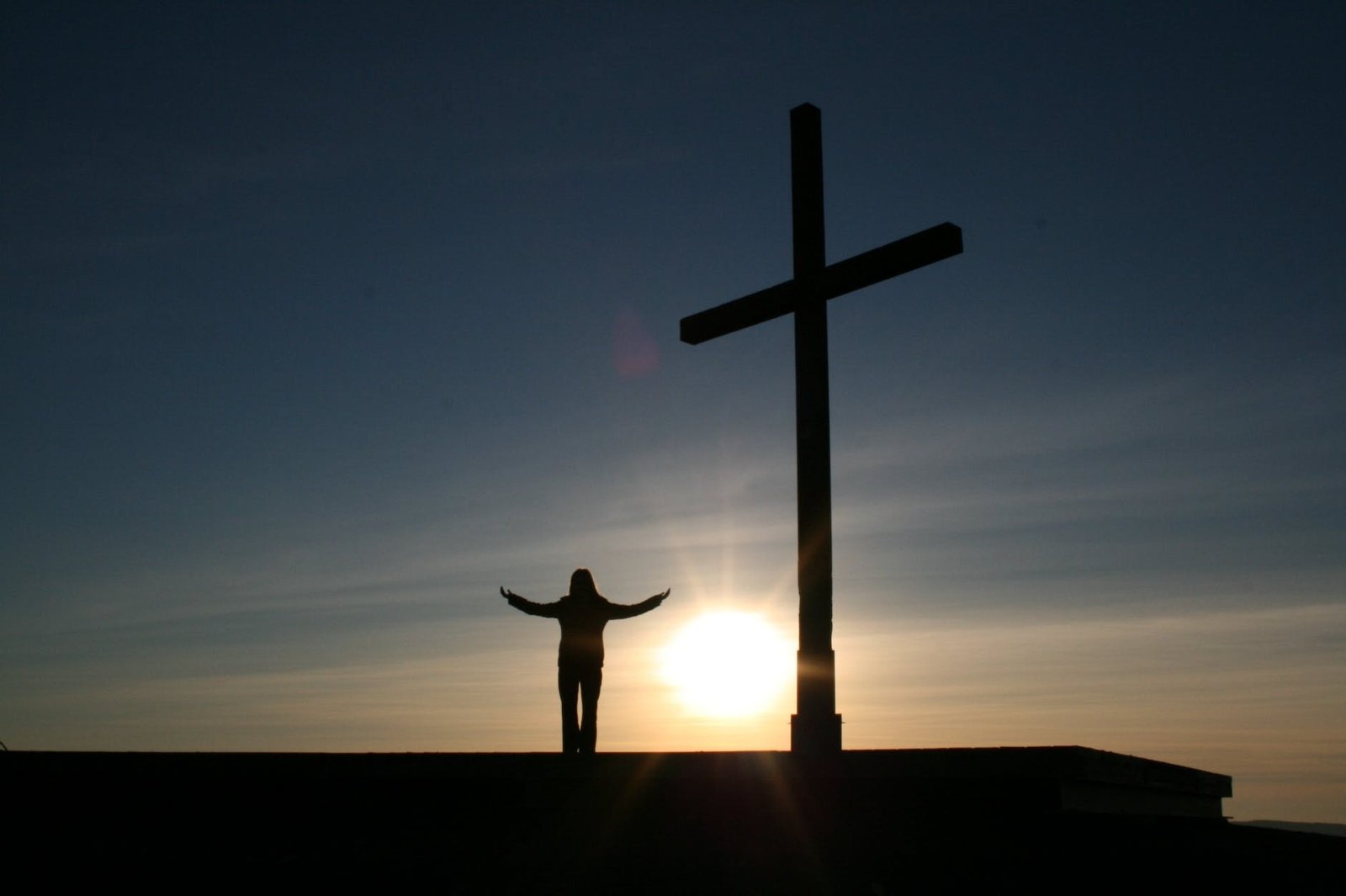 silhouette of person standing beside cross during sunset