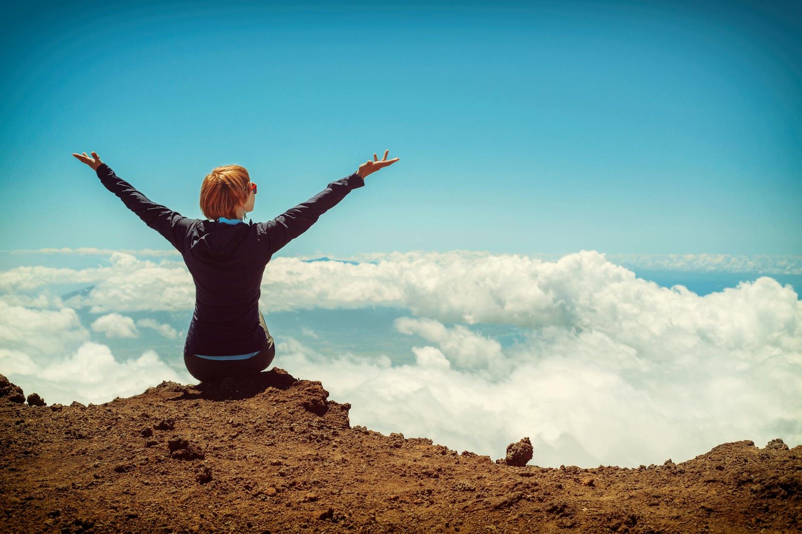 person sitting on cliff raising up both hands