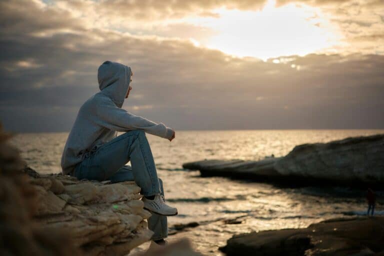 man in hoodie sitting on rock on sea shore at sunset